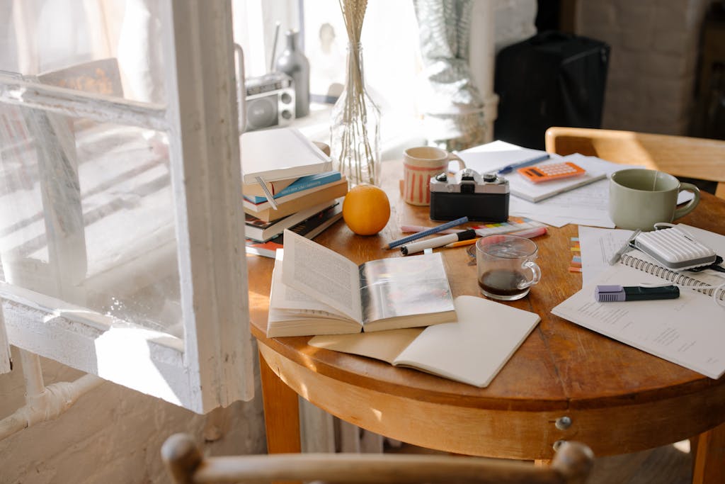 A cluttered study desk with open books, coffee, and sunlight streaming through a window.