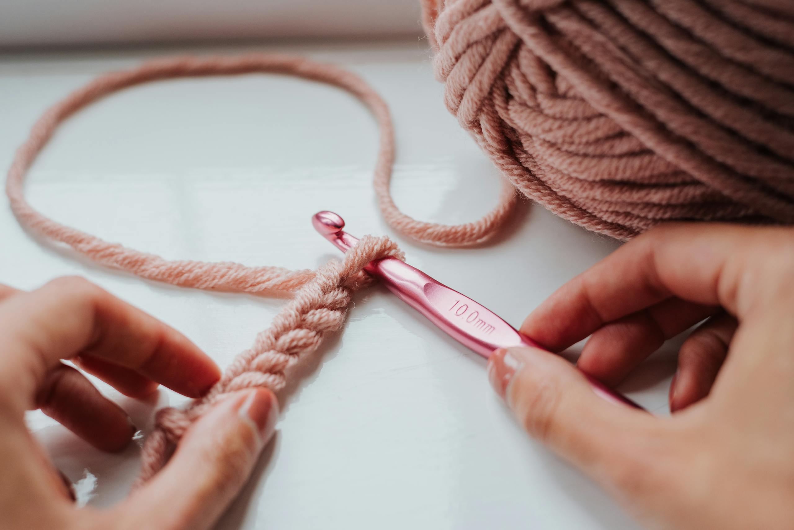 Hands crocheting with pink yarn and a 10mm hook on a cozy windowsill.